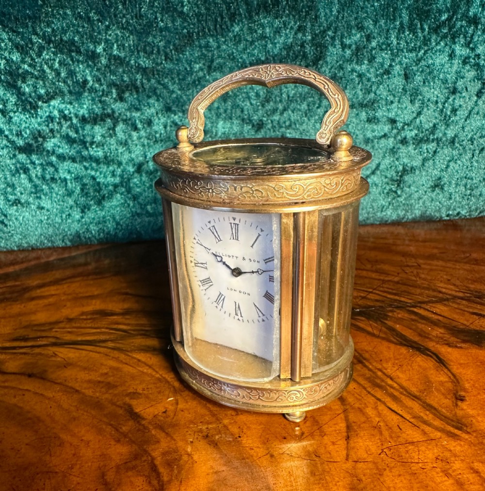A miniature brass carriage clock featuring engraved scrollwork borders, an overhead handle, curved glass sides, and a white dial with Roman numerals signed Elliott & Son London. View VI
