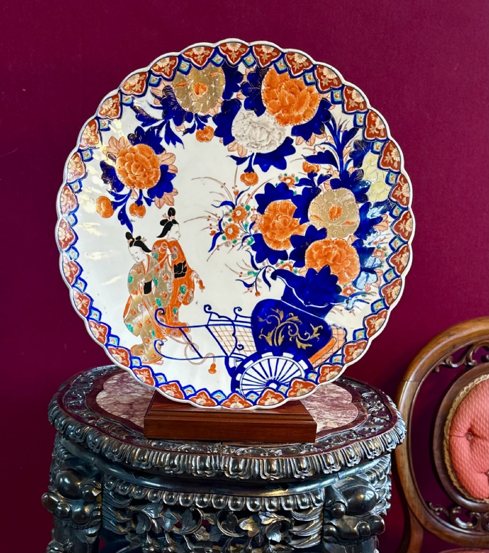 A circular Japanese Imari porcelain charger decorated with hand-painted human figures, floral arrangements, and a scalloped border in cobalt blue, rust red, and gold.