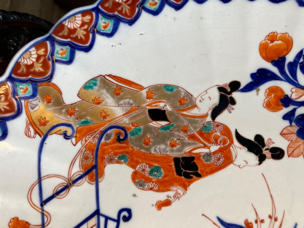 A circular Japanese Imari porcelain charger decorated with hand-painted human figures, floral arrangements, and a scalloped border in cobalt blue, rust red, and gold. View II