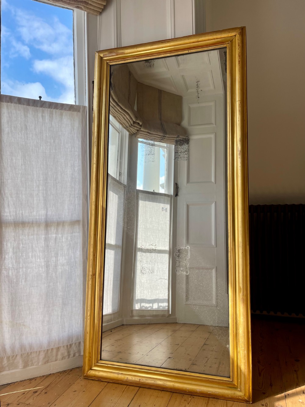 A tall antique French leaner mirror with an ornate gilded wooden frame resting on a wooden floor.