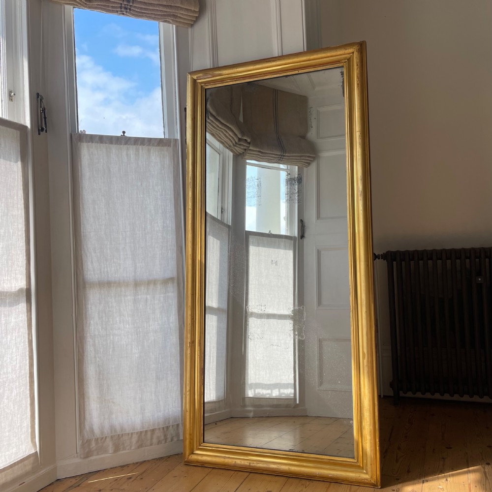 A tall antique French leaner mirror with an ornate gilded wooden frame resting on a wooden floor. View II