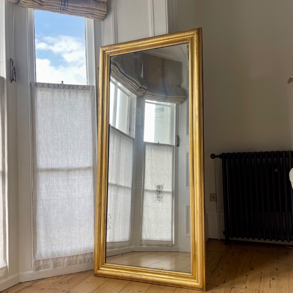 A tall antique French leaner mirror with an ornate gilded wooden frame resting on a wooden floor. View VI