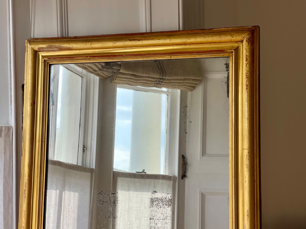 A tall antique French leaner mirror with an ornate gilded wooden frame resting on a wooden floor. View VIII