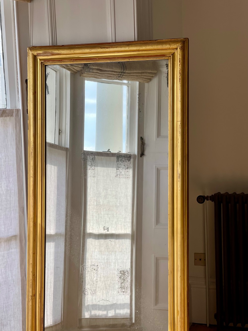 A tall antique French leaner mirror with an ornate gilded wooden frame resting on a wooden floor. View IX