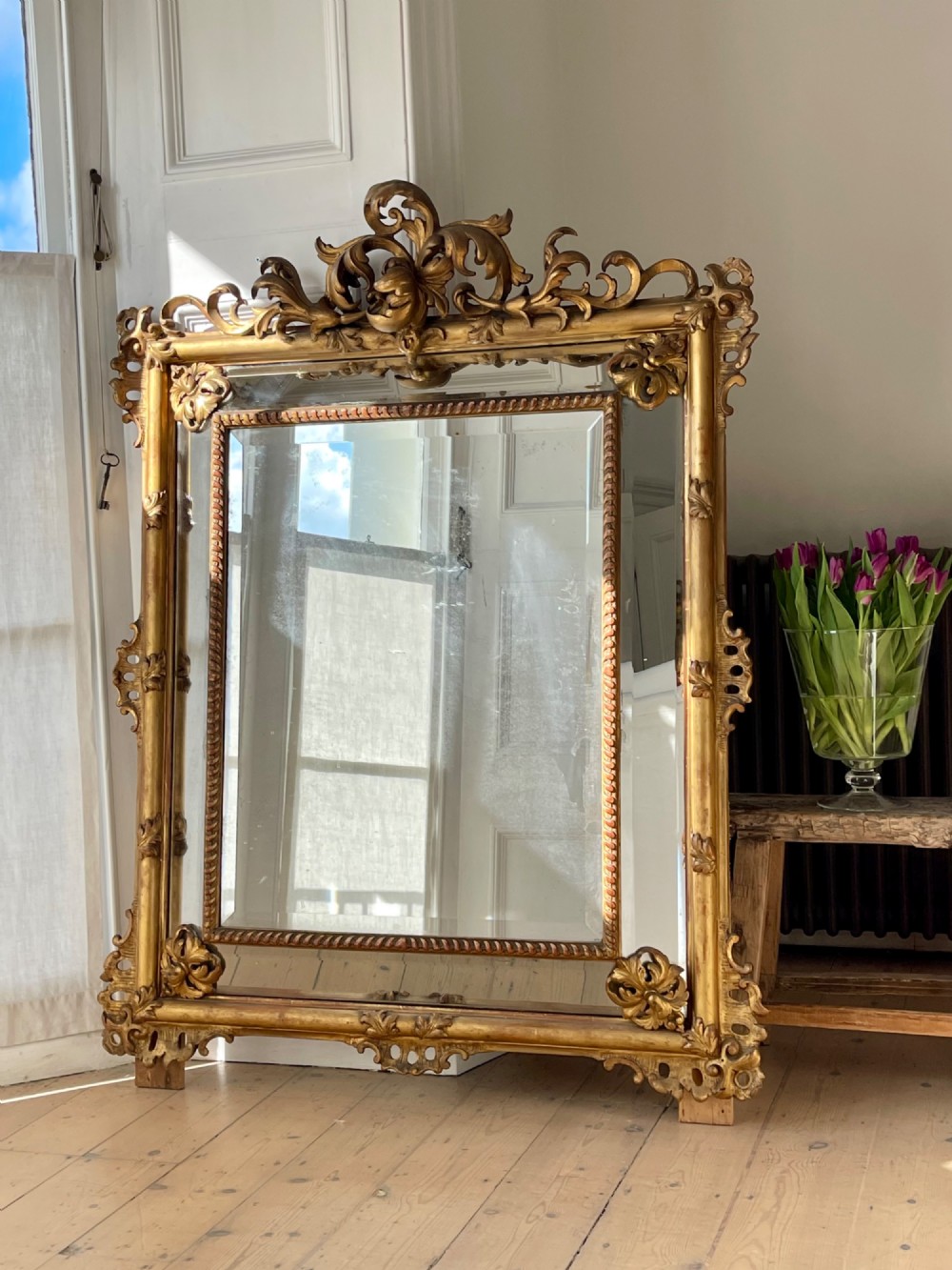 A large antique French giltwood wall mirror featuring carved acanthus cresting, corner flourishes, and bevelled mercury glass.