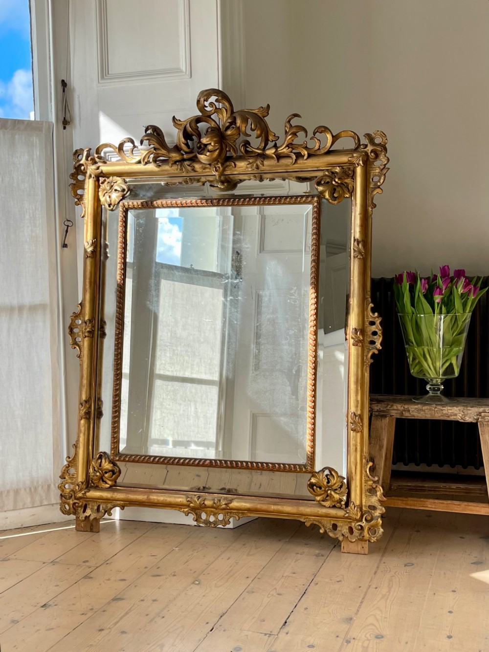 A large antique French giltwood wall mirror featuring carved acanthus cresting, corner flourishes, and bevelled mercury glass. View VI