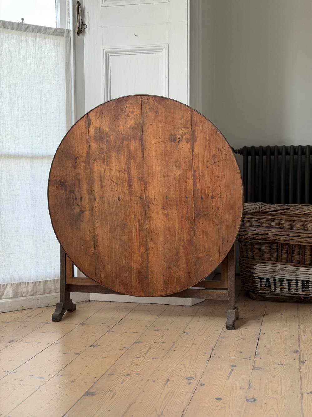 An antique French wooden vendange table featuring a circular plank top shown tilted vertically on a wooden trestle frame base. View II