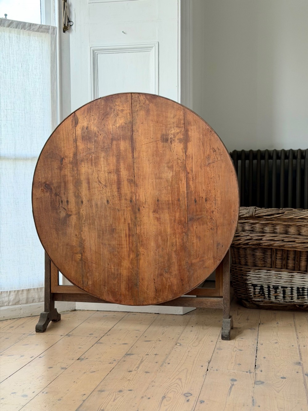 An antique French wooden vendange table featuring a circular plank top shown tilted vertically on a wooden trestle frame base. View III