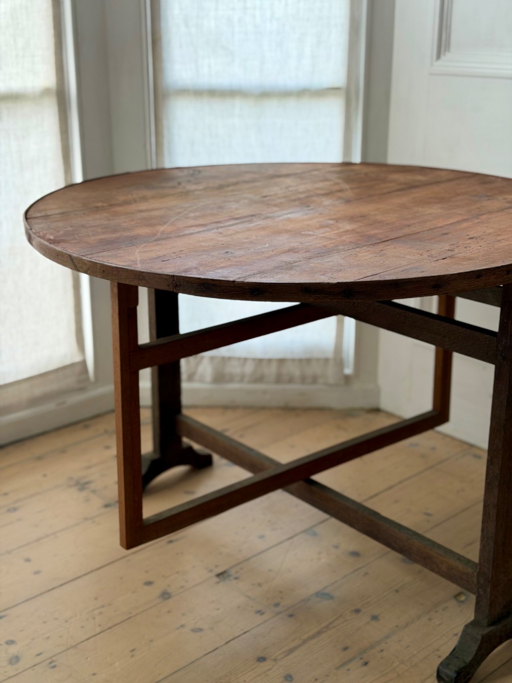 An antique French wooden vendange table featuring a circular plank top shown tilted vertically on a wooden trestle frame base. View X