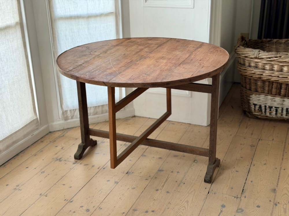 An antique French wooden vendange table featuring a circular plank top shown tilted vertically on a wooden trestle frame base. View XII