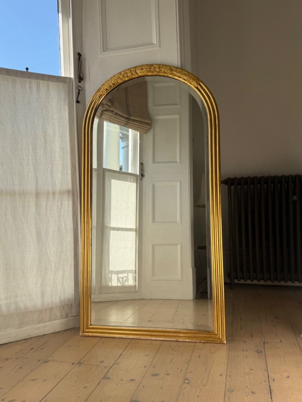 An arched tall mirror featuring a gilded gold frame with fluted side moulding and a carved floral crest. View II