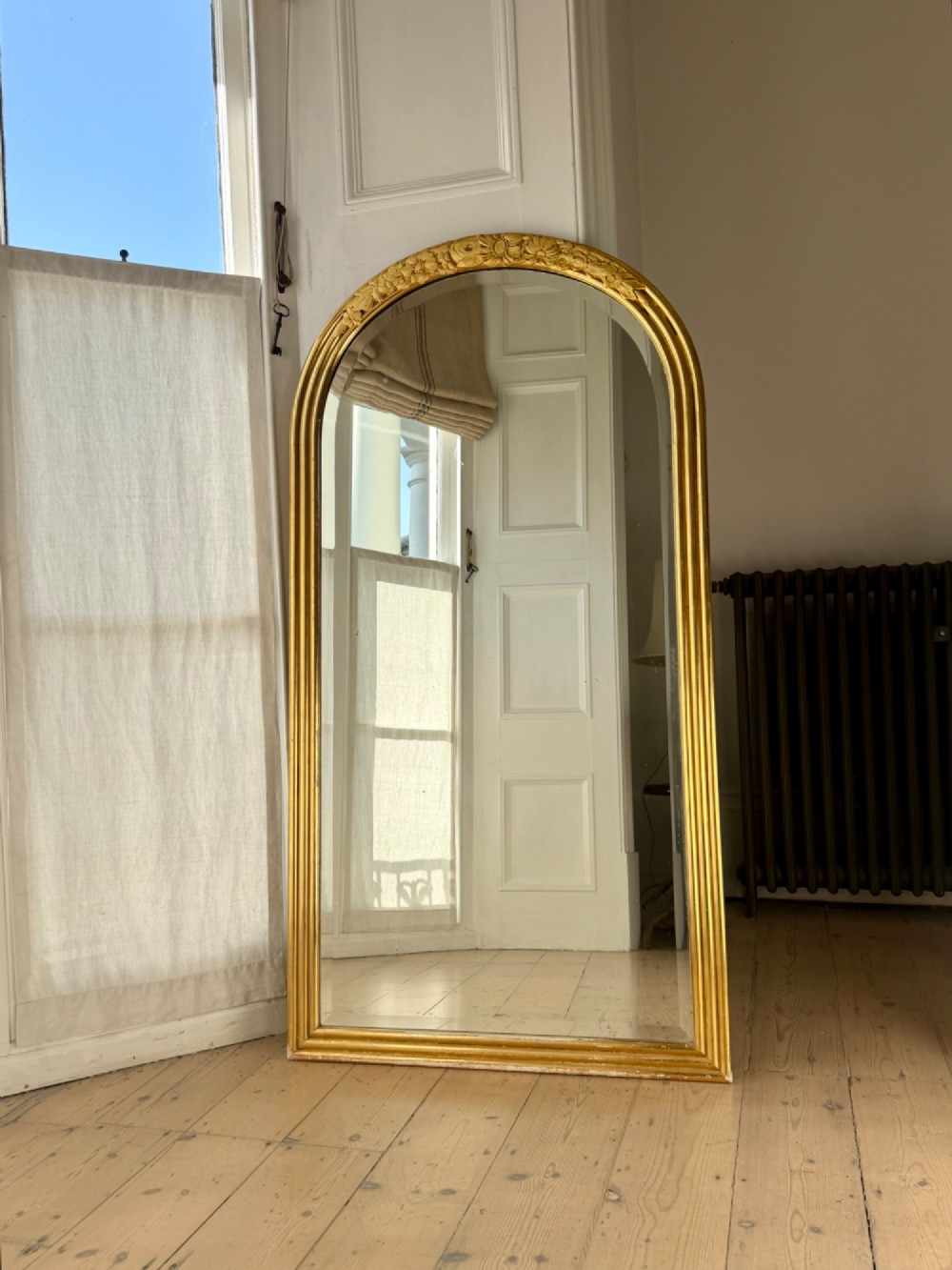 An arched tall mirror featuring a gilded gold frame with fluted side moulding and a carved floral crest.