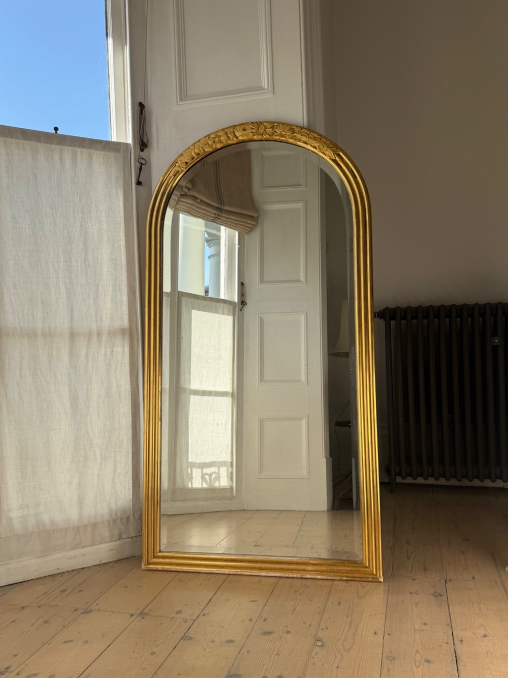 An arched tall mirror featuring a gilded gold frame with fluted side moulding and a carved floral crest. View VII