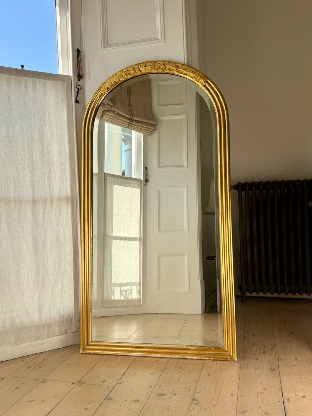 An arched tall mirror featuring a gilded gold frame with fluted side moulding and a carved floral crest. View VIII