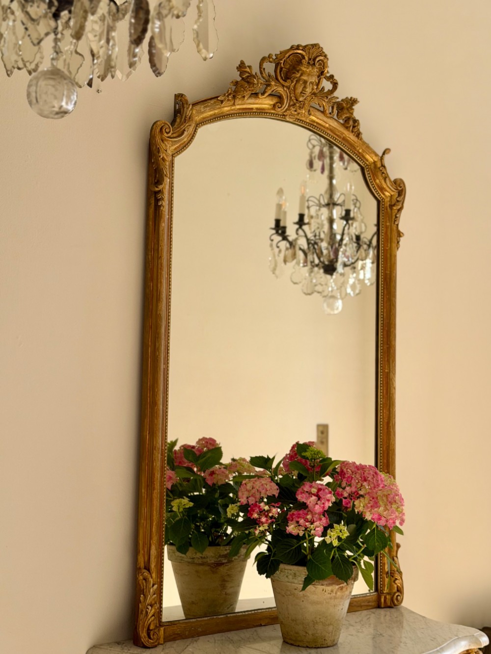 A tall arched antique French wall mirror featuring a gold leaf frame with a carved crest and beaded inner border.