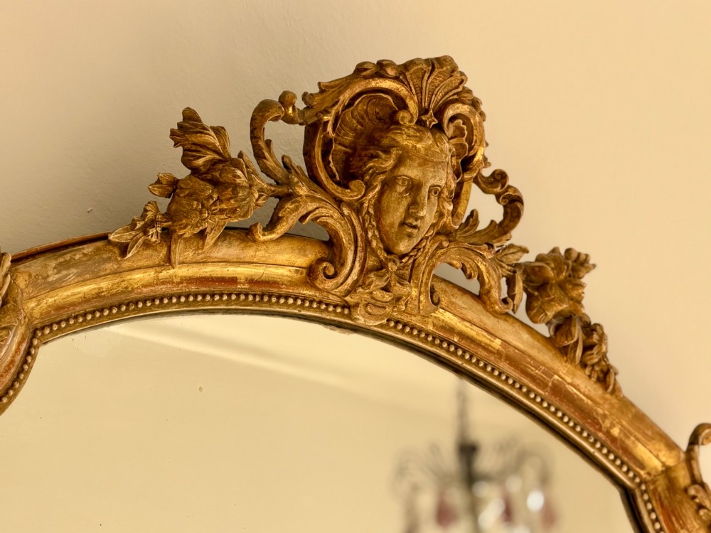 A tall arched antique French wall mirror featuring a gold leaf frame with a carved crest and beaded inner border. View VI