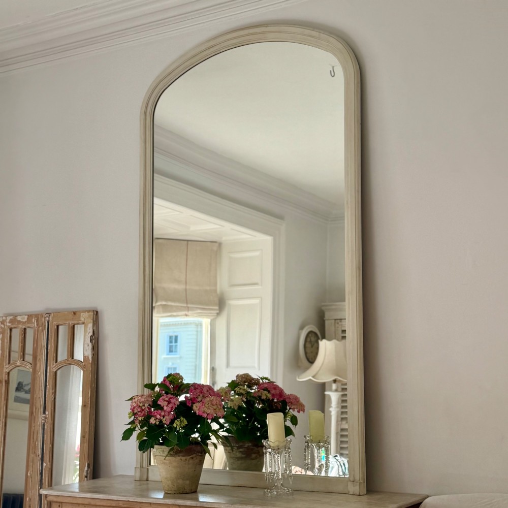 A tall arched mirror featuring a cream painted wooden frame resting on a surface.