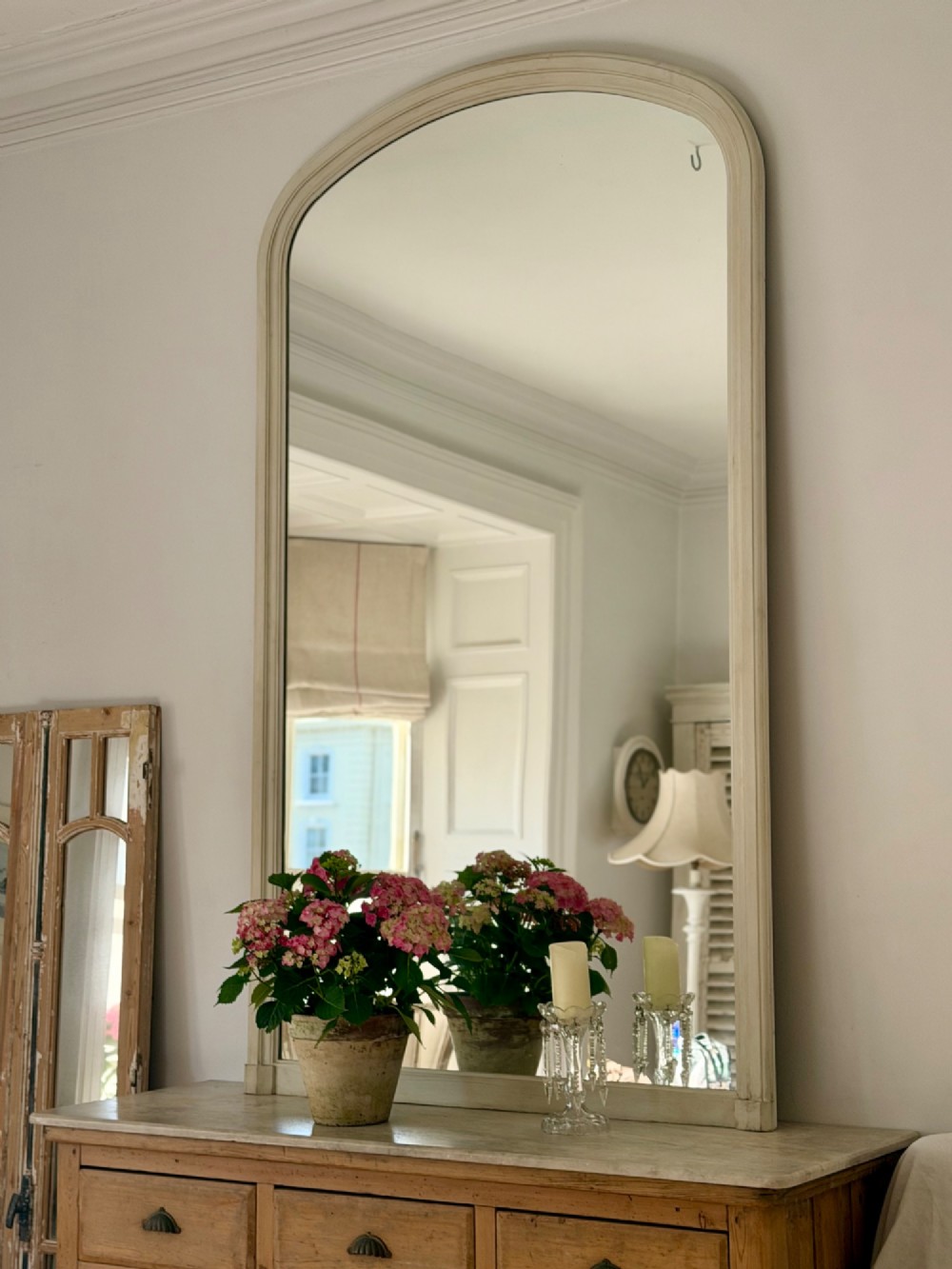A tall arched mirror featuring a cream painted wooden frame resting on a surface. View II