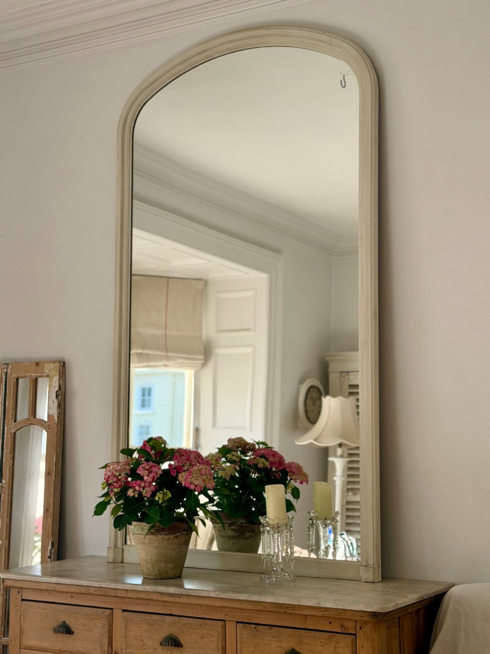 A tall arched mirror featuring a cream painted wooden frame resting on a surface. View III