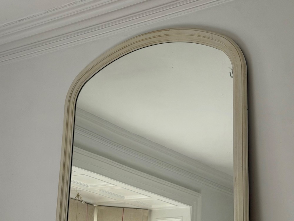 A tall arched mirror featuring a cream painted wooden frame resting on a surface. View VI