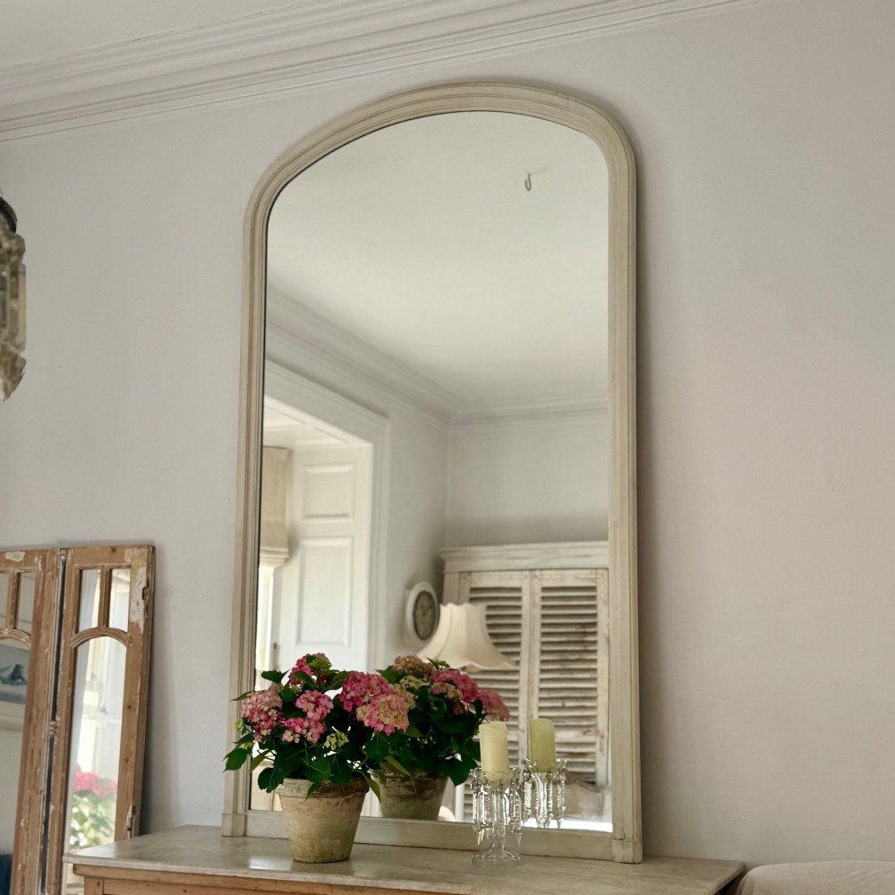 A tall arched mirror featuring a cream painted wooden frame resting on a surface. View IX