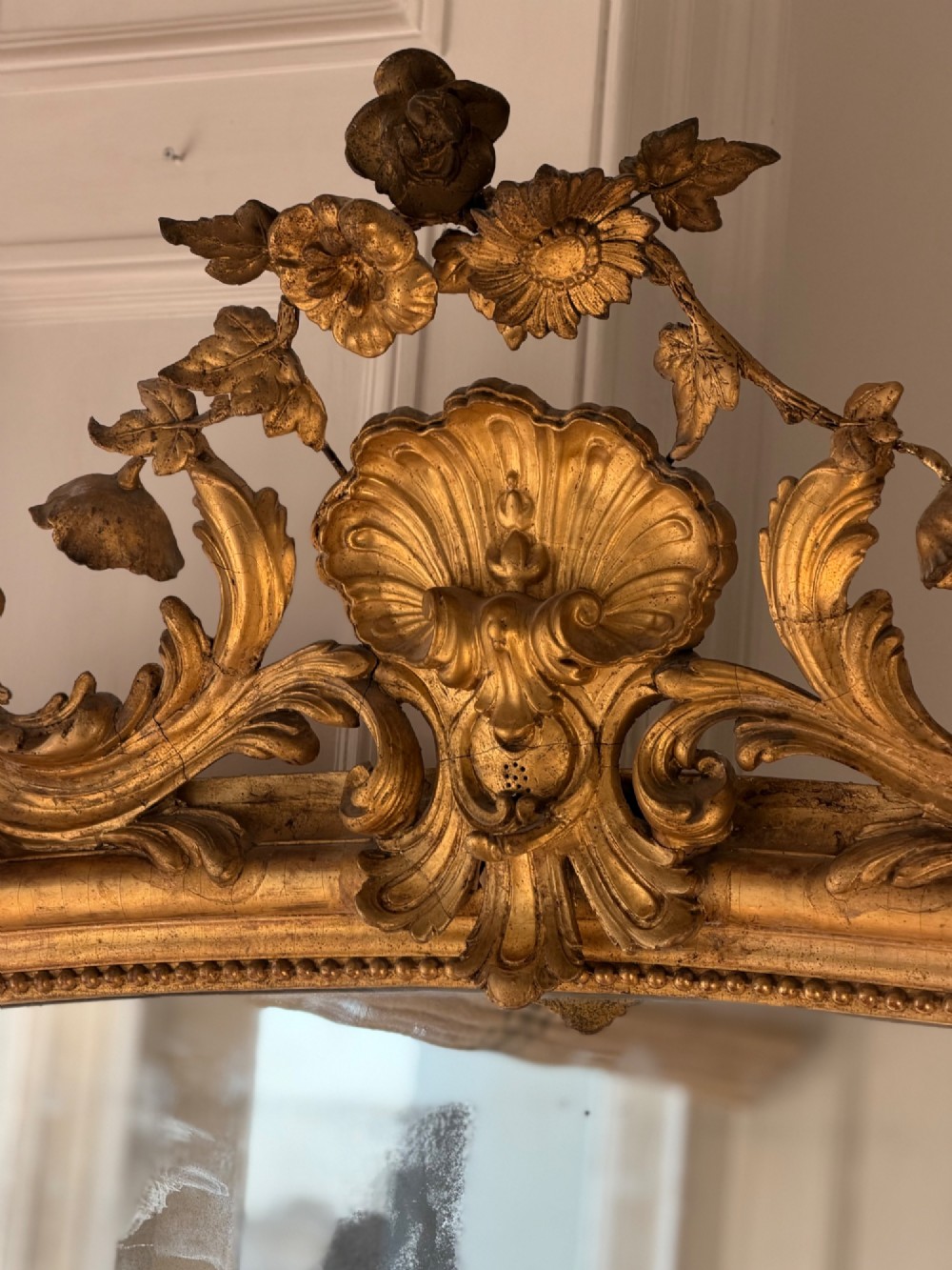 A large 18th century French giltwood mirror featuring an ornate floral shell crest and visible foxing on the mercury glass plate. View IV