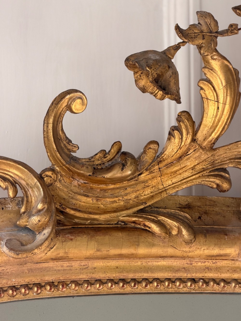 A large 18th century French giltwood mirror featuring an ornate floral shell crest and visible foxing on the mercury glass plate. View IX