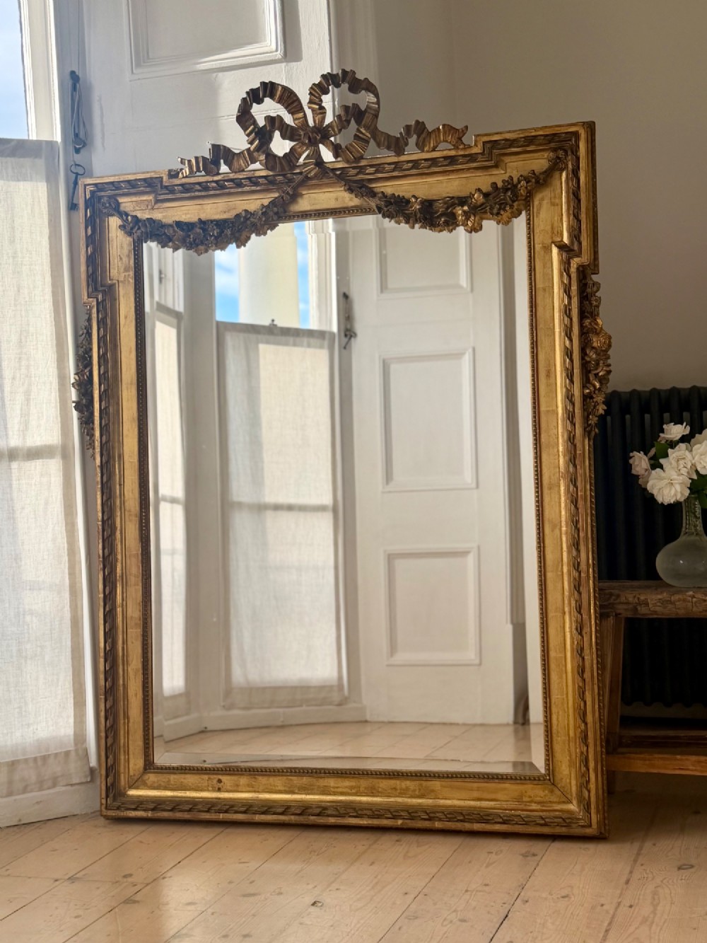 A rectangular French Louis XVI style giltwood mirror featuring a carved ribbon cresting, draped floral swags, and a bevelled glass mirror plate.