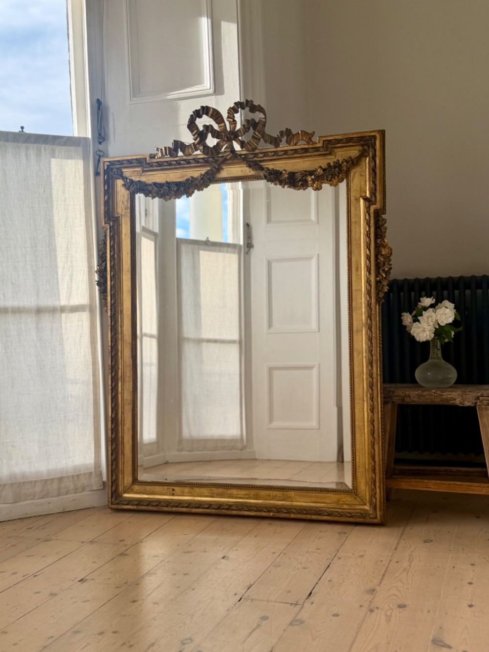 A rectangular French Louis XVI style giltwood mirror featuring a carved ribbon cresting, draped floral swags, and a bevelled glass mirror plate. View II