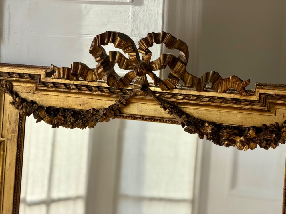 A rectangular French Louis XVI style giltwood mirror featuring a carved ribbon cresting, draped floral swags, and a bevelled glass mirror plate. View V