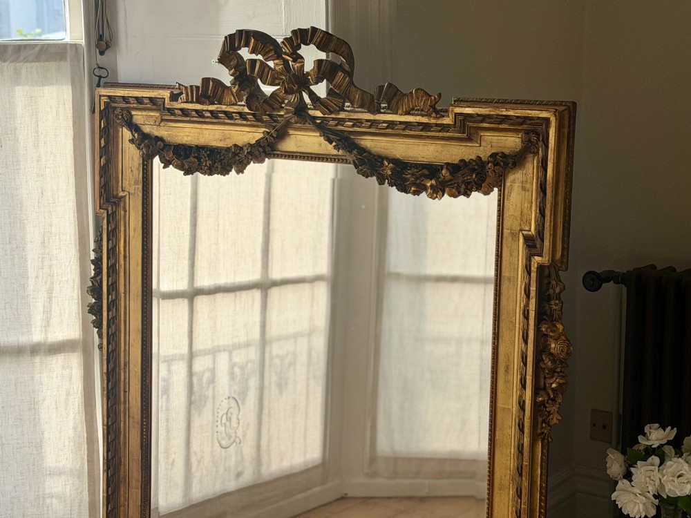 A rectangular French Louis XVI style giltwood mirror featuring a carved ribbon cresting, draped floral swags, and a bevelled glass mirror plate. View VI