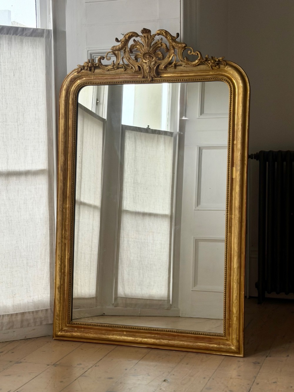 A tall French Louis Philippe mirror featuring a gold leaf frame with rounded upper shoulders, pearl bead moulding, and a decorative carved crest.