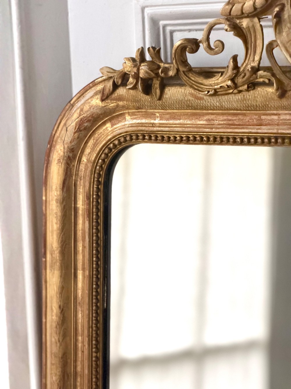 A tall French Louis Philippe mirror featuring a gold leaf frame with rounded upper shoulders, pearl bead moulding, and a decorative carved crest. View V
