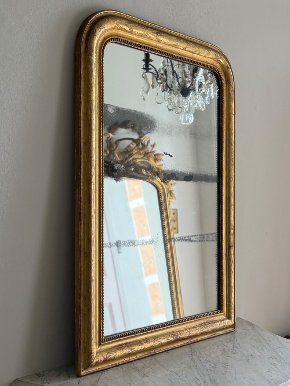 A rectangular antique French gilt mirror featuring rounded upper corners, an engraved gold leaf frame with beaded inner moulding, and foxed mercury glass.