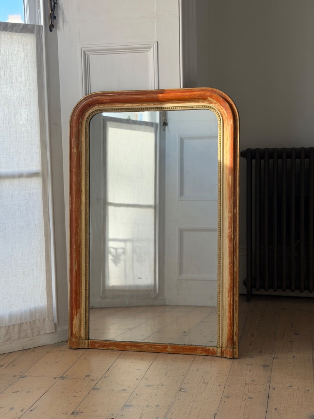 A French Louis Philippe mirror featuring a rounded-top frame with worn gold leaf finish exposing the red clay bole beneath, holding a mercury glass plate. View II