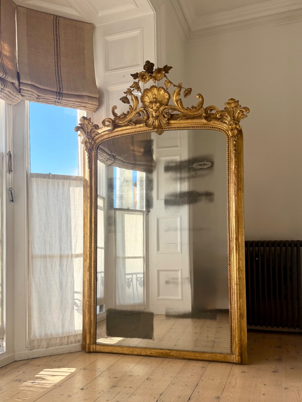 A large 18th century French giltwood mirror featuring an ornate floral shell crest and visible foxing on the mercury glass plate. View XIII