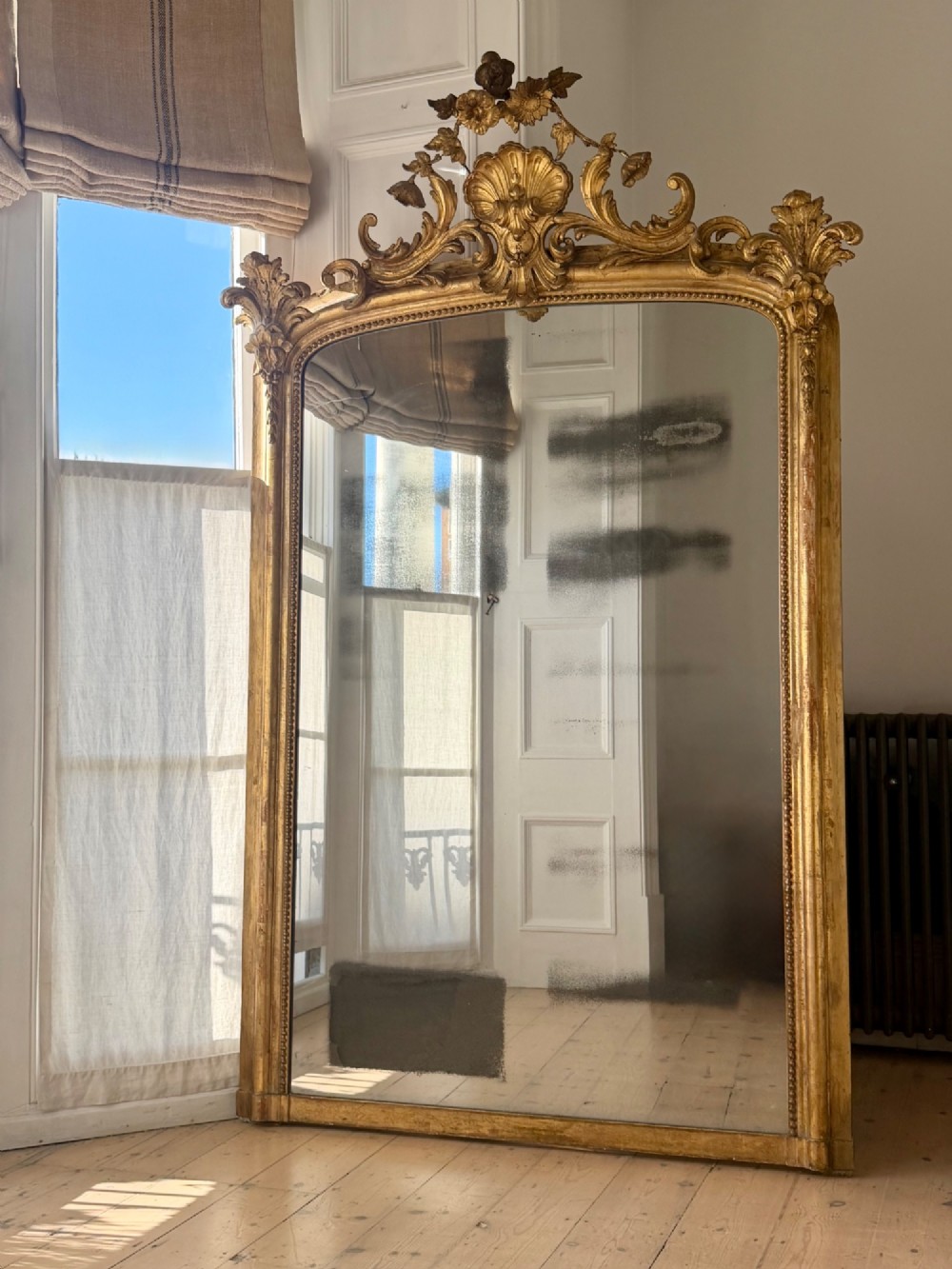 A large 18th century French giltwood mirror featuring an ornate floral shell crest and visible foxing on the mercury glass plate.
