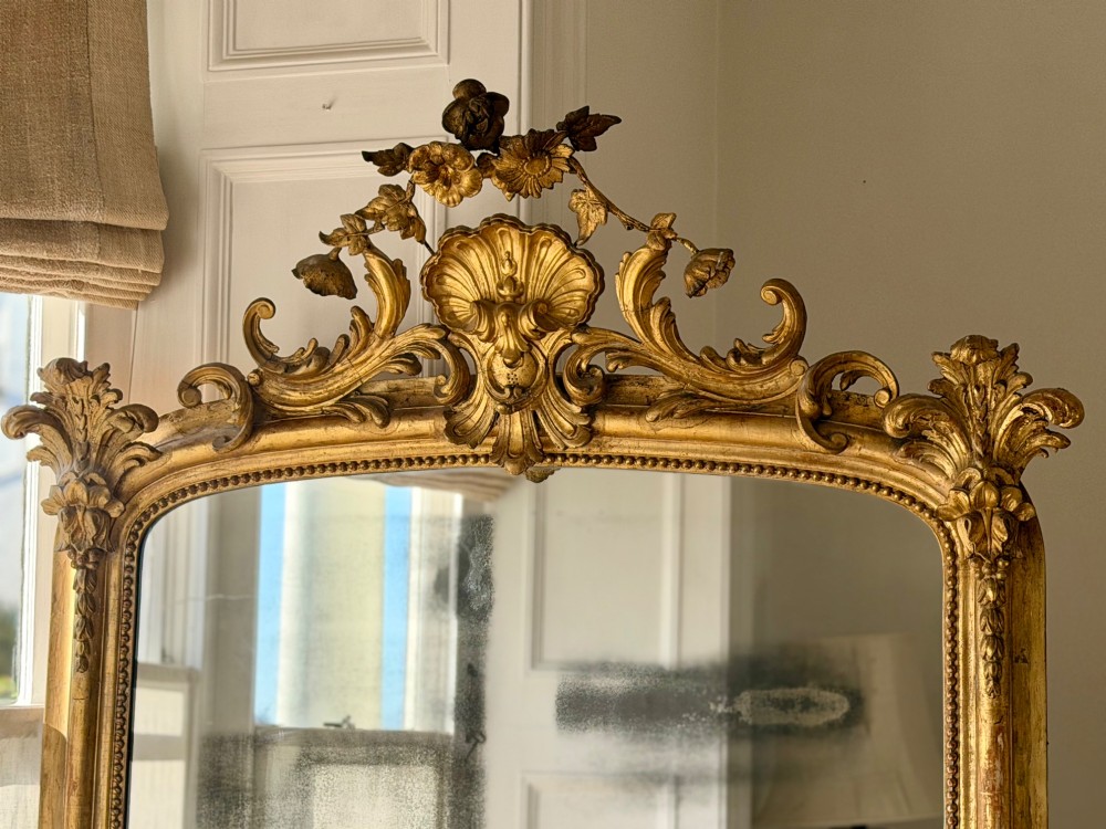 A large 18th century French giltwood mirror featuring an ornate floral shell crest and visible foxing on the mercury glass plate. View XIV