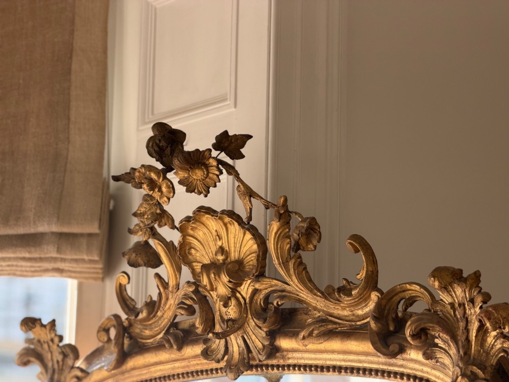 A large 18th century French giltwood mirror featuring an ornate floral shell crest and visible foxing on the mercury glass plate. View XV
