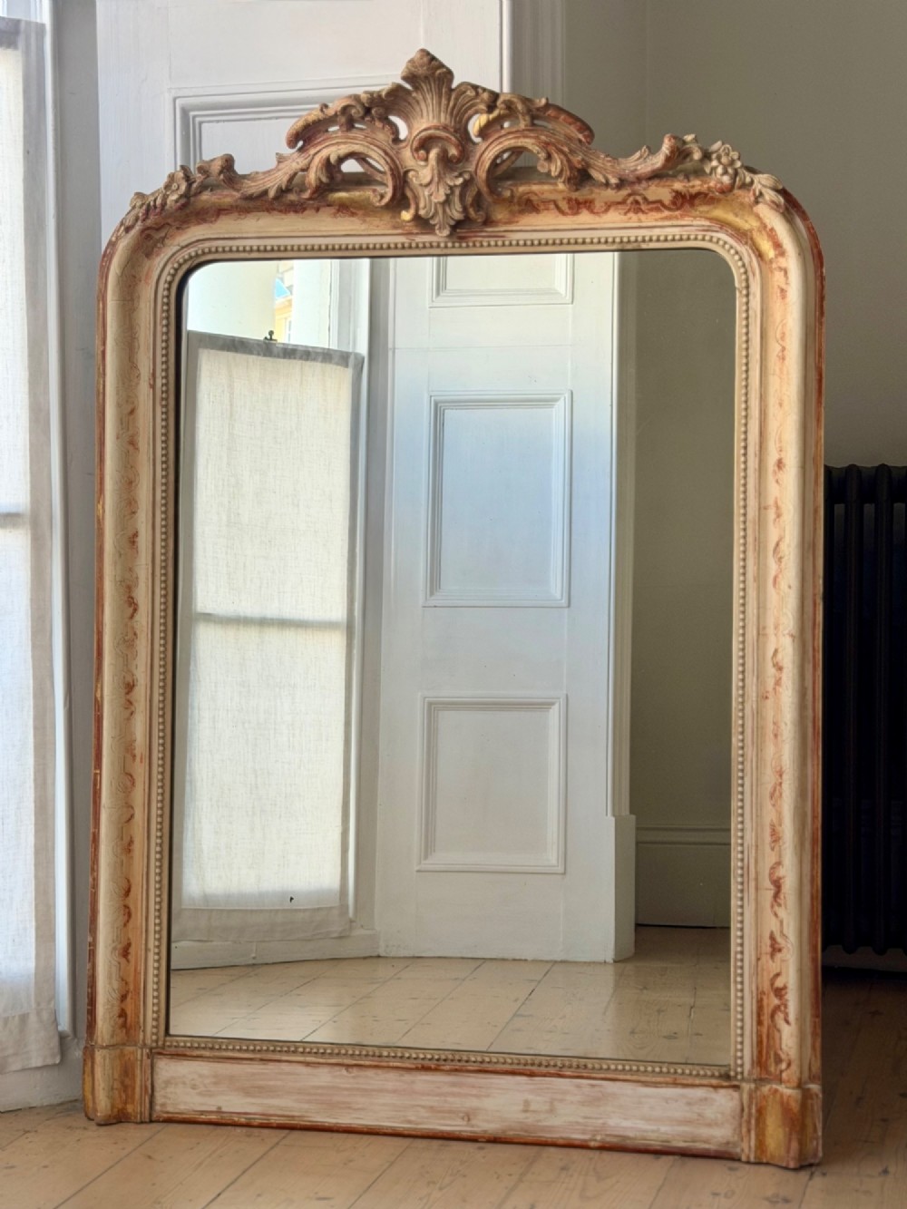 An antique French mirror featuring an arched top frame with a carved cresting and distressed off-white gesso finish.