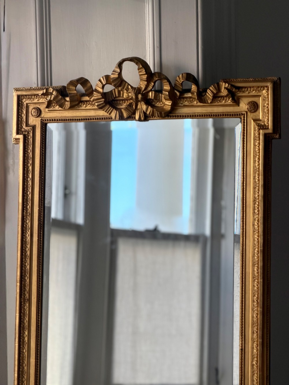 A tall rectangular gold giltwood full-length mirror featuring a carved bow ribbon cresting, beaded inner frame, and bevelled glass plate. View VI