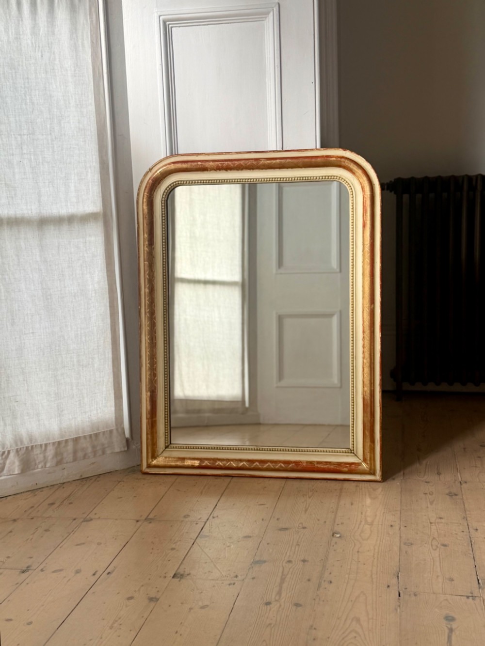 An antique French wall mirror with a rounded arched top, distressed gold leaf and cream gesso frame featuring incised geometric patterns and beaded inner edge.
