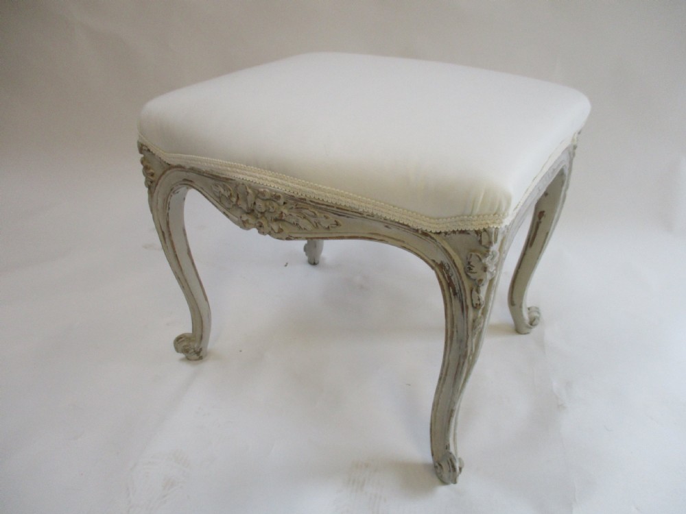 A French Louis XV style rectangular stool with hand-carved floral aprons, distressed grey cabriole legs, and a cream upholstered top.