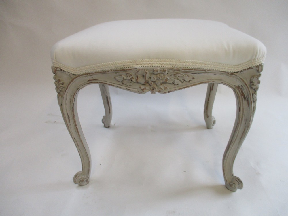 A French Louis XV style rectangular stool with hand-carved floral aprons, distressed grey cabriole legs, and a cream upholstered top. View II