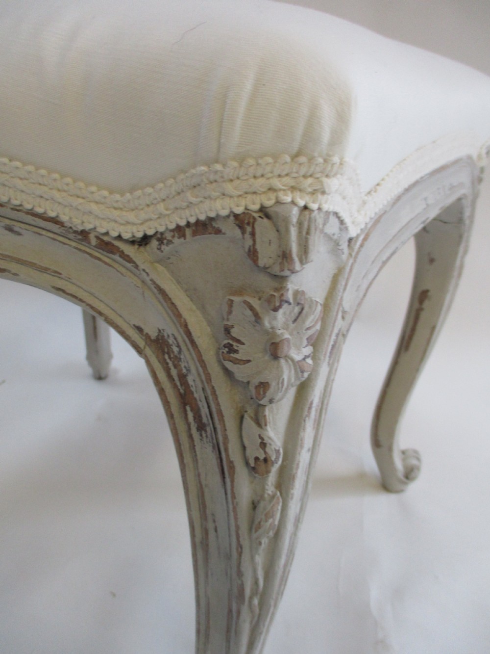 A French Louis XV style rectangular stool with hand-carved floral aprons, distressed grey cabriole legs, and a cream upholstered top. View IV