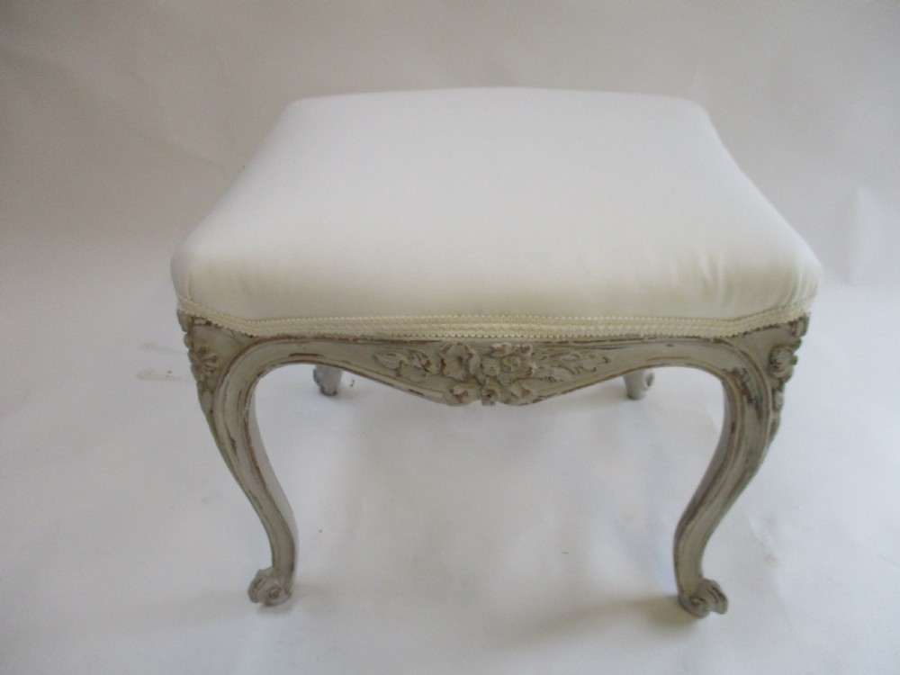 A French Louis XV style rectangular stool with hand-carved floral aprons, distressed grey cabriole legs, and a cream upholstered top. View VI