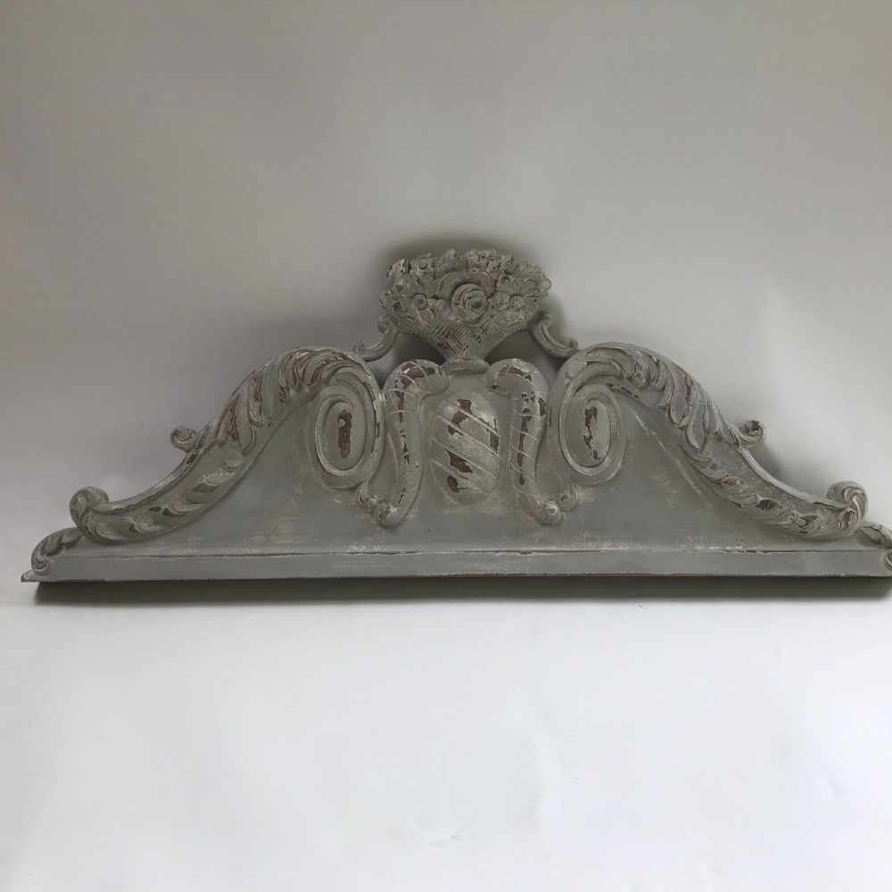 A carved wooden architectural pelmet featuring central scroll motifs and a distressed grey painted finish.