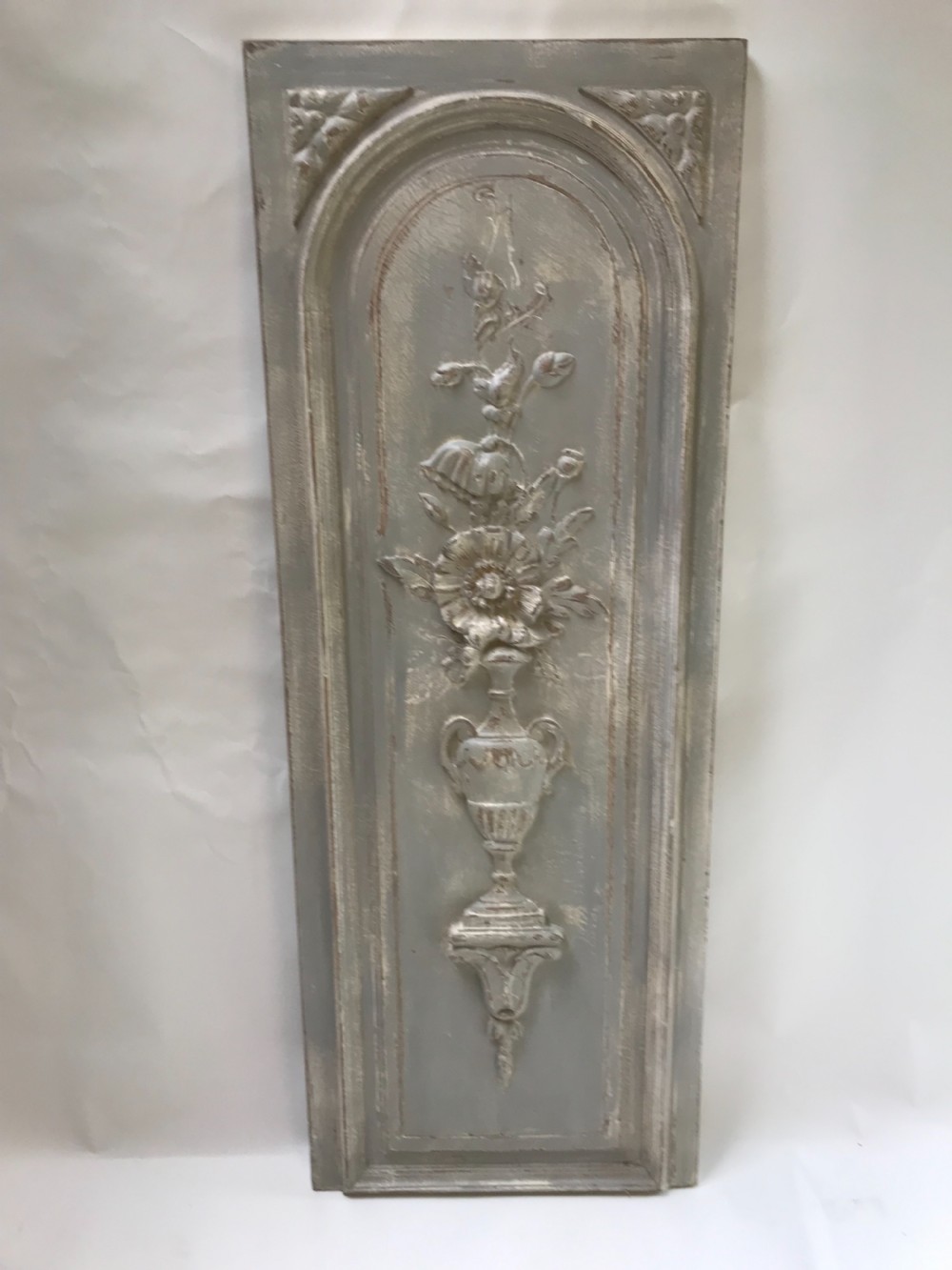 A vertical painted grey wood panel featuring an arched frame encircling a hand-carved classical urn and floral relief.