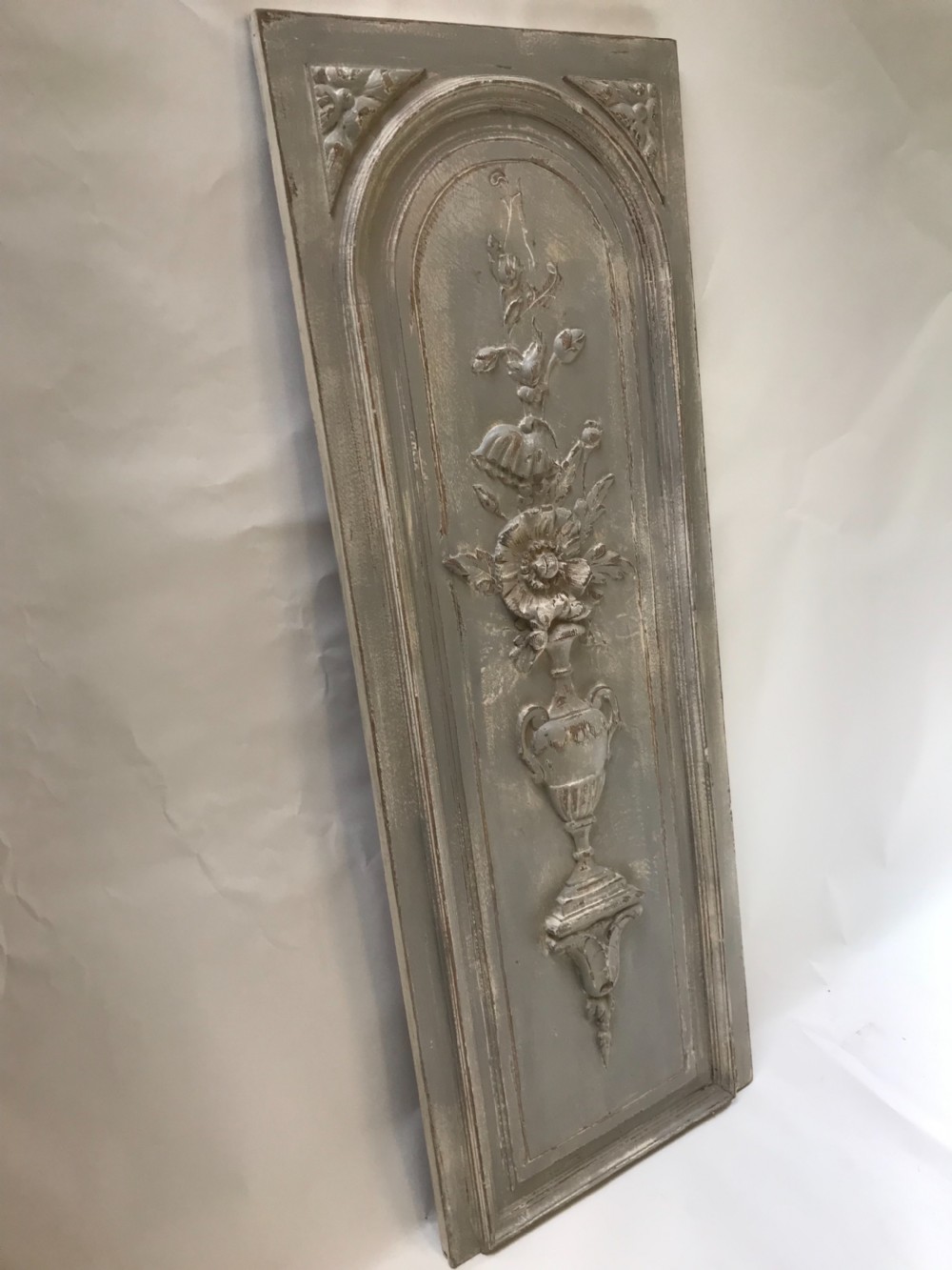 A vertical painted grey wood panel featuring an arched frame encircling a hand-carved classical urn and floral relief. View III
