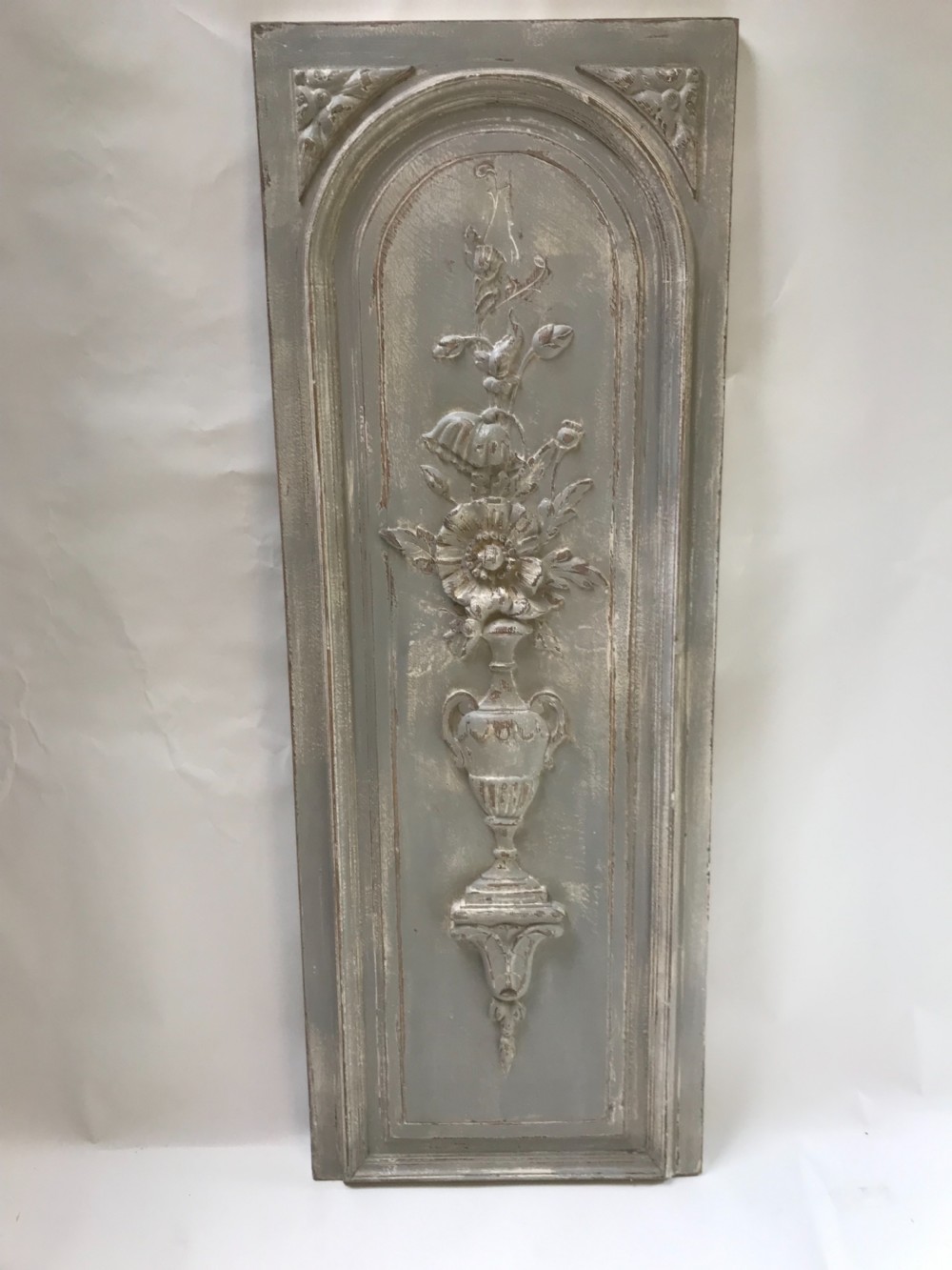 A vertical painted grey wood panel featuring an arched frame encircling a hand-carved classical urn and floral relief. View IV